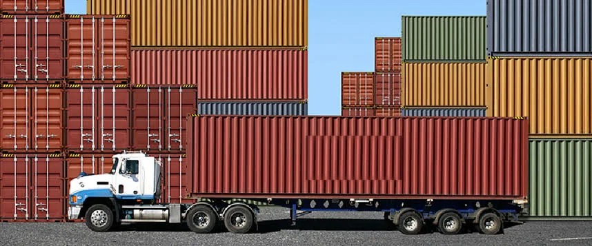 A truck hauling a plain shipping container through a port yard, with stacks of containers in several colours rising behind it.