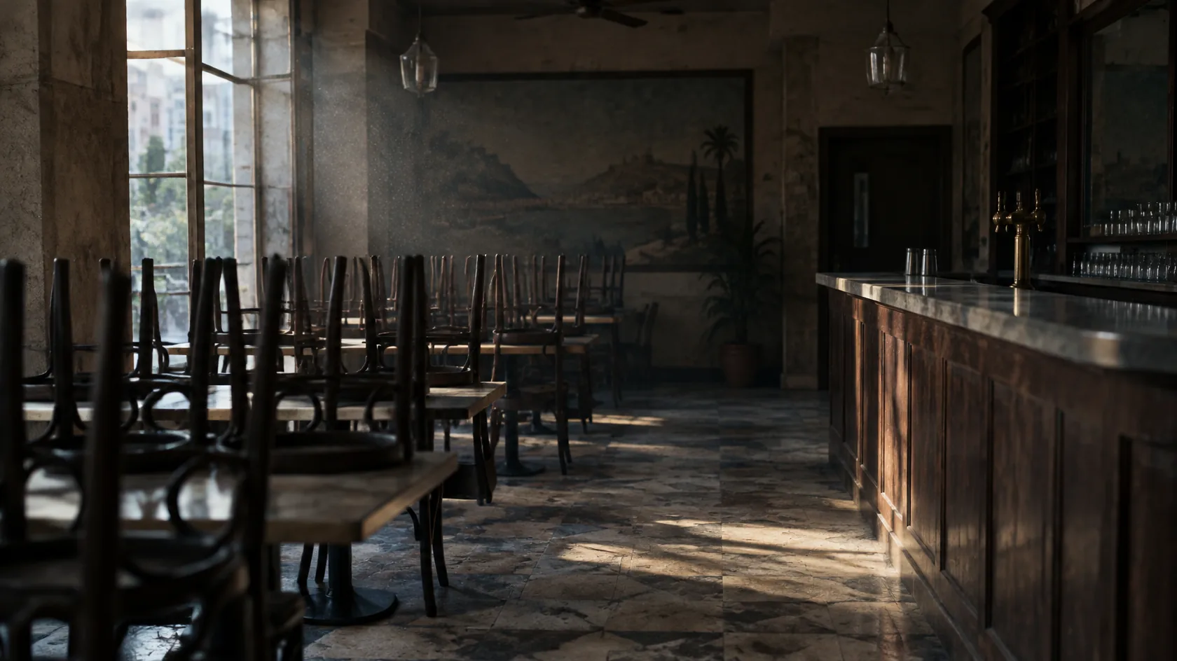 The empty dining room of a closed restaurant, chairs stacked upside down on the tables and the bar counter bare, with late afternoon light falling through tall windows onto the tiled floor.